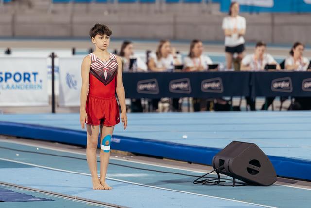 Young male gymnast stands barefoot on the floor mat, composed and focused, wearing a red leotard with knee support.