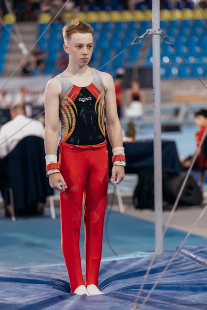 Young male gymnast stands composed and focused near high bar apparatus, wearing red pants and agiva leotard.