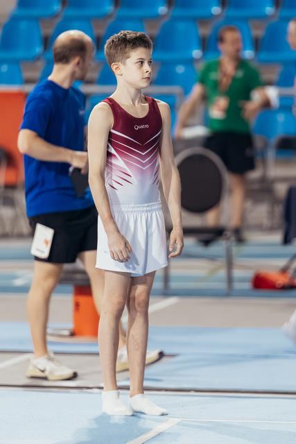 Young male gymnast in red and white Quatro leotard stands attentively on the floor, calm and focused, awaiting his turn.