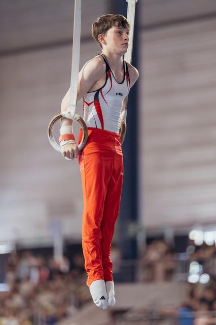 Young male gymnast performing on still rings, holding a static position with intense focus in a competition hall.