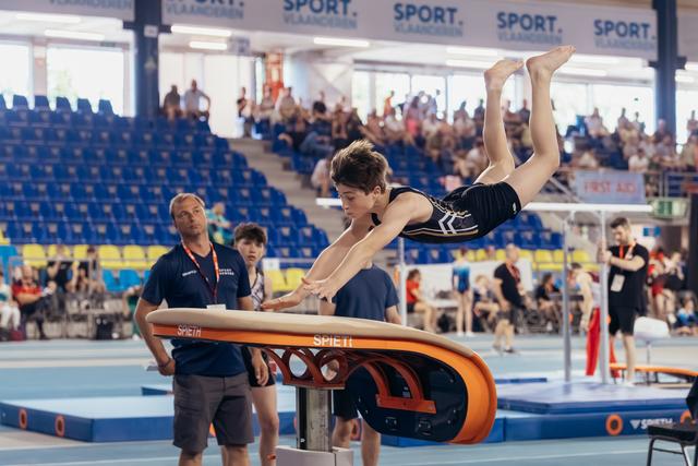 Young male gymnast soars horizontal over a Spieth vault table, arms reaching down, legs extended, at an indoor gymnastics meet.