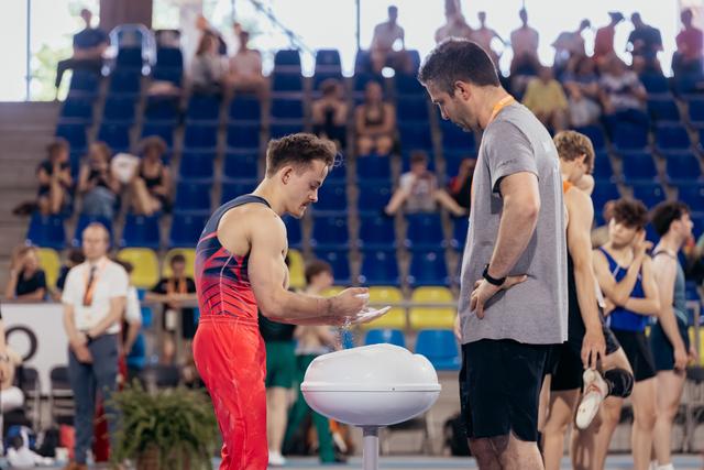 Young gymnast applies chalk to hands at a bowl while his coach watches attentively in a busy competition arena.