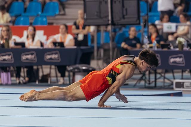 Male gymnast in red uniform executes a dramatic floor dive, body parallel to the mat at a gymnastics competition.