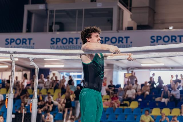 Young male gymnast grips parallel bars with intense grimace, wearing green and black leotard, crowd watching in background.