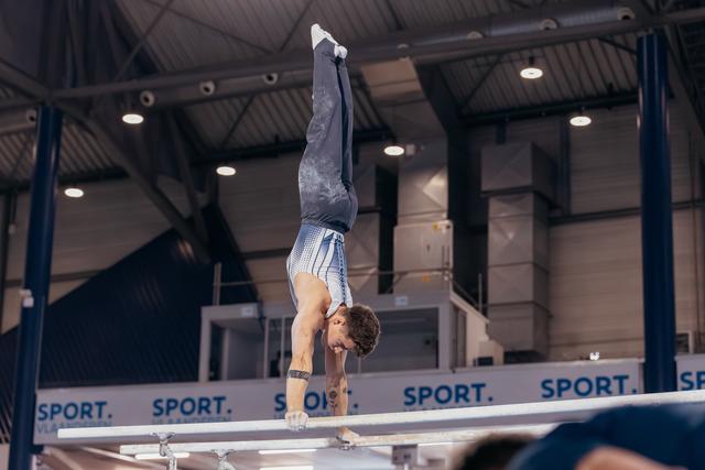 Male gymnast performs a perfect handstand on parallel bars, body fully extended, in an indoor sports hall.