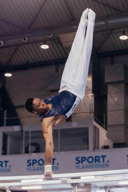 Male gymnast holds a powerful one-arm handstand on parallel bars, legs raised high, at Sport Vlaanderen event.