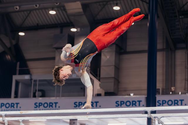 Young male gymnast in red and black leotard performs inverted release on parallel bars at indoor sports hall.