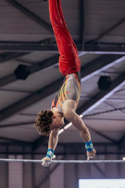 Male gymnast inverts powerfully on the high bar, legs raised in red pants, showing intense concentration and strength.