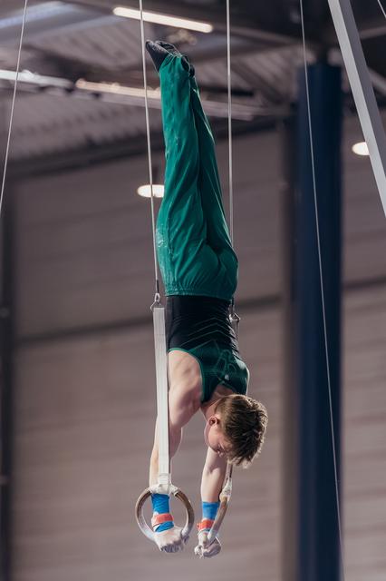 Young male gymnast performs an inverted hang on still rings, body fully extended upside down in a sports hall.
