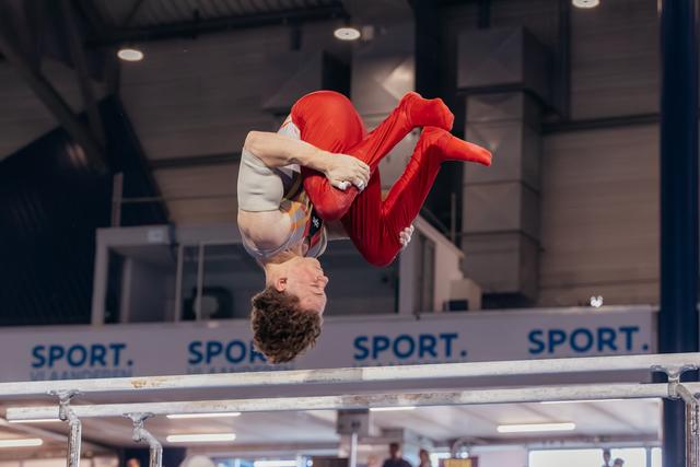 Young male gymnast upside-down in a tucked somersault above parallel bars, smiling mid-flight in an indoor sports hall.