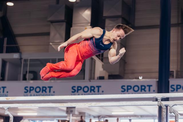 Male gymnast soars above parallel bars in red and navy uniform, body tucked, face intense with effort.