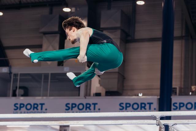 Male gymnast mid-air above parallel bars, legs tucked and extended, focused expression, in teal uniform at indoor arena.