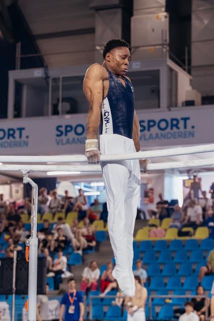 Male gymnast grips parallel bars with intense focus, muscular arms engaged, competing before a packed arena crowd.