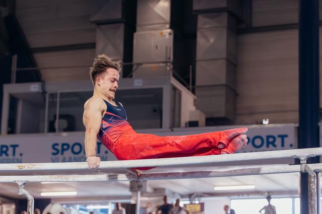 Male gymnast performs a straddle L-sit on parallel bars, jaw clenched in intense concentration, at an indoor sports hall.