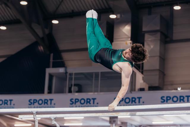 Male gymnast holds a dramatic inverted position on parallel bars, green leotard, intense focus, indoor arena.
