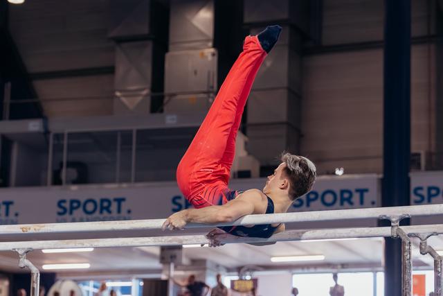 Young male gymnast performs a straddle-L hold on parallel bars, legs raised high, focused expression in a sports hall.