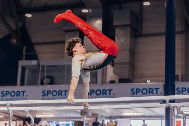 Male gymnast performs an inverted pike hold on parallel bars, legs raised in red uniform, intense concentration indoors.