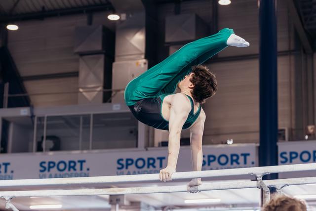 Young male gymnast performs a pike hold on parallel bars, body folded with intense focus, at indoor sports venue.