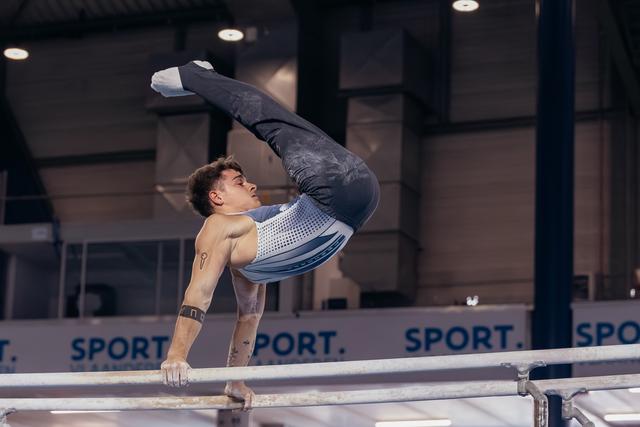 Male gymnast performs a dramatic pike hold on parallel bars, body arched with legs extended upward in a competition hall.