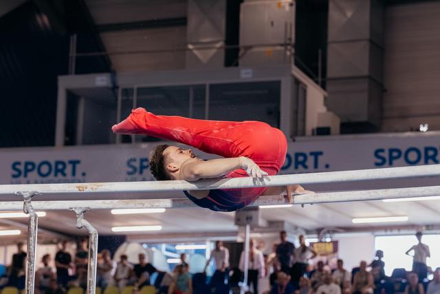 Male gymnast in red performs a tucked pike hold on parallel bars, body folded with focused expression, crowd watching.