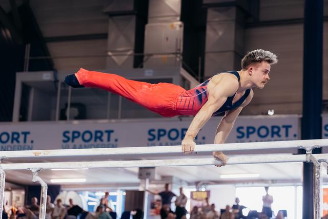 Male gymnast holds a planche on parallel bars, body horizontal, expression intense and determined at indoor sports event.