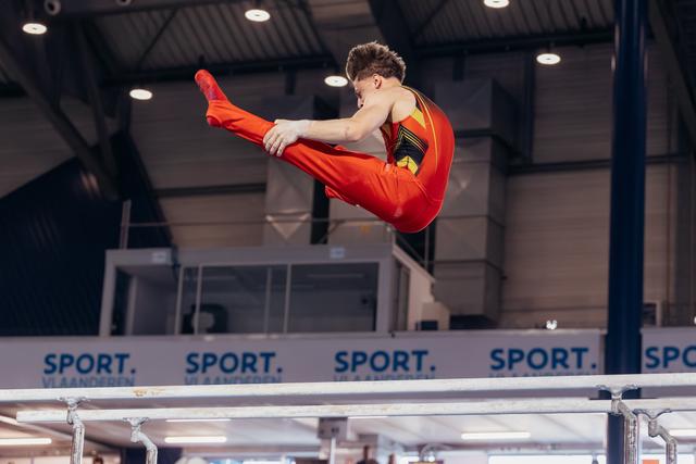 Male gymnast in red suit performs a dynamic straddle jump above parallel bars, legs fully extended, intense focus.