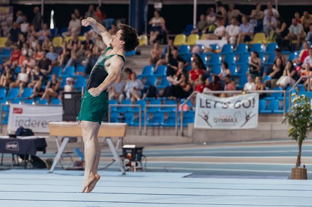 Male gymnast leaps with fist raised in triumph, green uniform, performing before a packed indoor arena crowd.