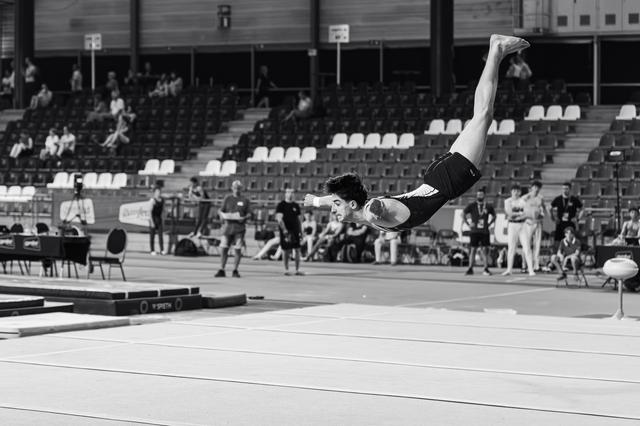 Male gymnast mid-backflip over the floor mat, body fully extended, during an indoor gymnastics competition.