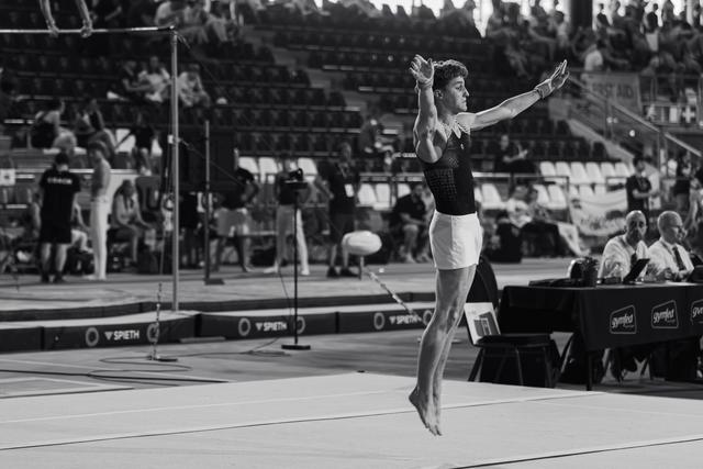 Male gymnast mid-air during floor exercise, arms gracefully extended, focused expression, judges seated nearby.