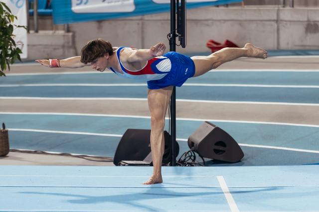Young male gymnast holds a perfect arabesque balance on one leg, arms extended, focused expression, on blue competition floor.