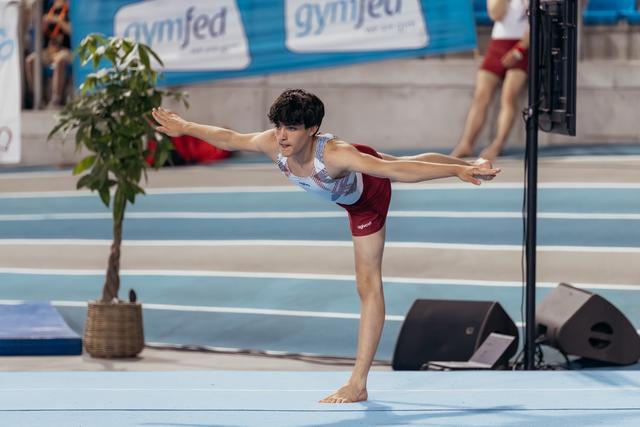 Young male gymnast holds a precise arabesque balance on the floor, arms outstretched, focused expression at a Gymfed event.