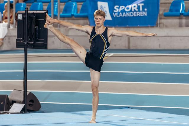 Young male gymnast holds a perfect standing split, arms outstretched, focused expression on blue indoor track.
