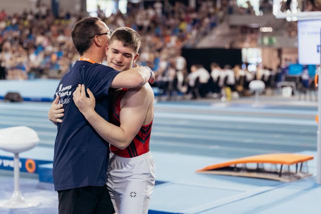 Young male gymnast hugs his coach on the competition floor, both sharing a warm, emotional moment of support.