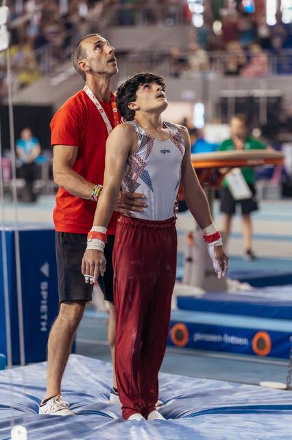 Young male gymnast and coach both gaze upward, coach steadying him with hands on shoulders in a supportive moment.