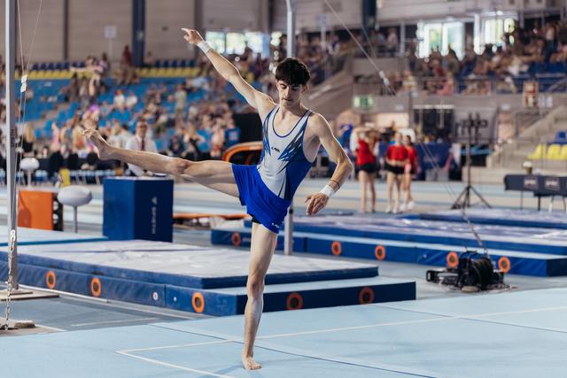 Young male gymnast performs a dynamic leg raise on the floor exercise, arms extended, focused expression in a busy arena.