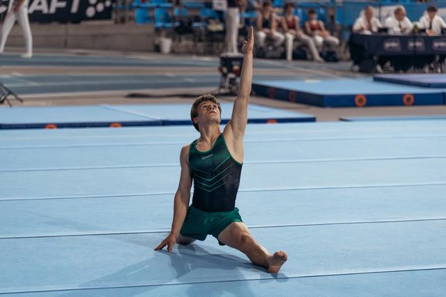 Young male gymnast in green leotard performs dramatic floor pose, arm raised skyward, head tilted back with intensity.