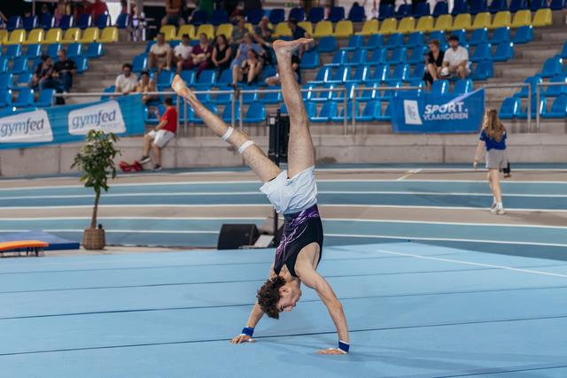 Young male gymnast performs a one-armed handstand on the blue floor, legs split wide, at a Gymfed indoor competition.