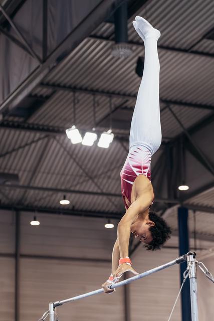 Male gymnast performs a perfect handstand on the horizontal bar, body fully extended, legs pointing skyward in an indoor arena.