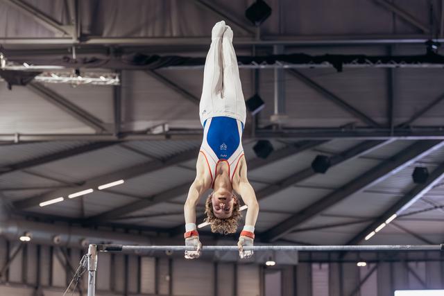 Young male gymnast holds a handstand on the high bar, legs straight up, wearing a blue and white leotard in an indoor arena.