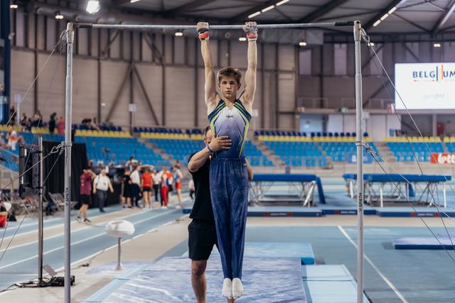 Young male gymnast hangs from high bar with focused expression while coach steadies him in Belgian sports hall.