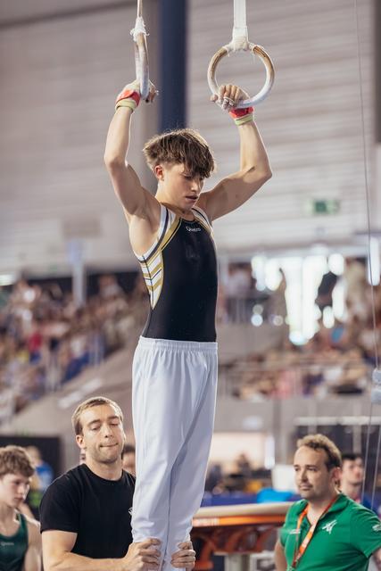 Young male gymnast hangs from still rings with intense focus, supported by coach below in a packed competition arena.