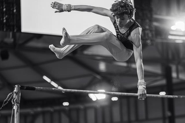 Young male gymnast mid-flight above the high bar, legs split, intense focus, wearing wrist guards in an indoor arena.