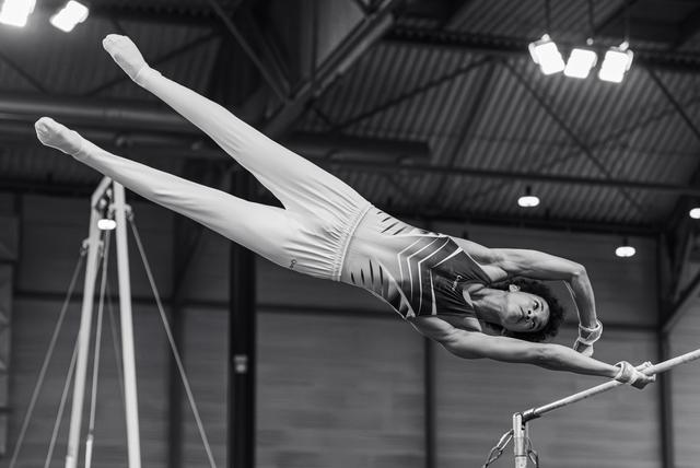Young gymnast performs a dramatic release move on the high bar, body fully extended in a swan dive position, indoor gym.