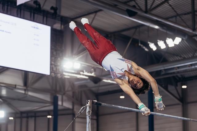 Male gymnast releases the high bar mid-flight, body fully extended and inverted, competing in an indoor gymnastics arena.