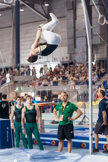 Gymnast performs an inverted flip mid-air while coach watches intently below, teammates observing in a packed competition arena.