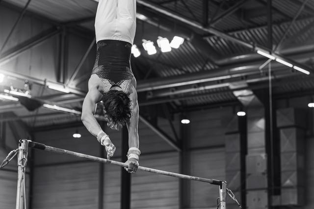 Male gymnast performs an inverted handstand on the high bar, muscles tensed, in a large indoor gymnastics hall.