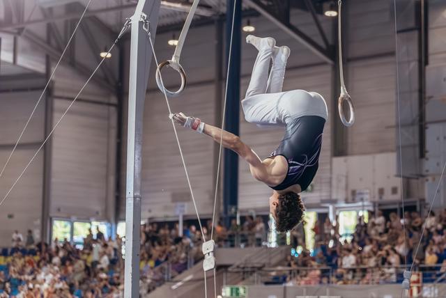 Young male gymnast hangs inverted on still rings, body arched with intense focus during a packed indoor gymnastics event.