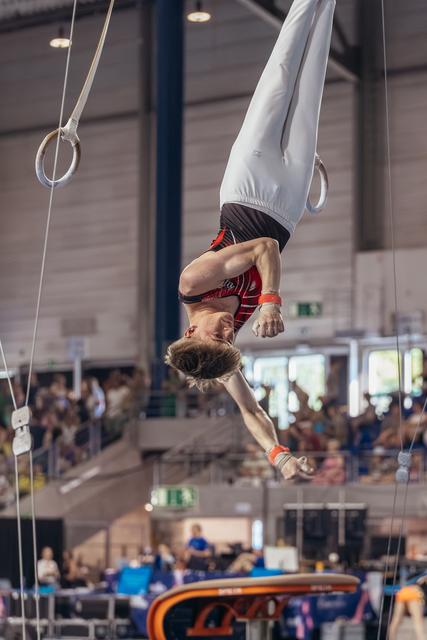 Male gymnast performs an inverted hang on still rings, body fully extended, during a packed indoor gymnastics competition.