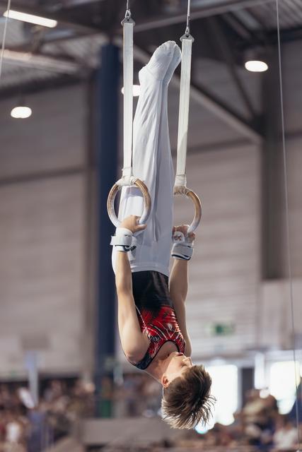 Young male gymnast performs an inverted hang on still rings, body fully extended, in a packed competition gymnasium.