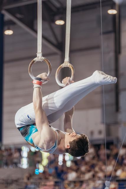 Young male gymnast performs an inverted tuck hold on still rings, showing intense concentration during a competition.